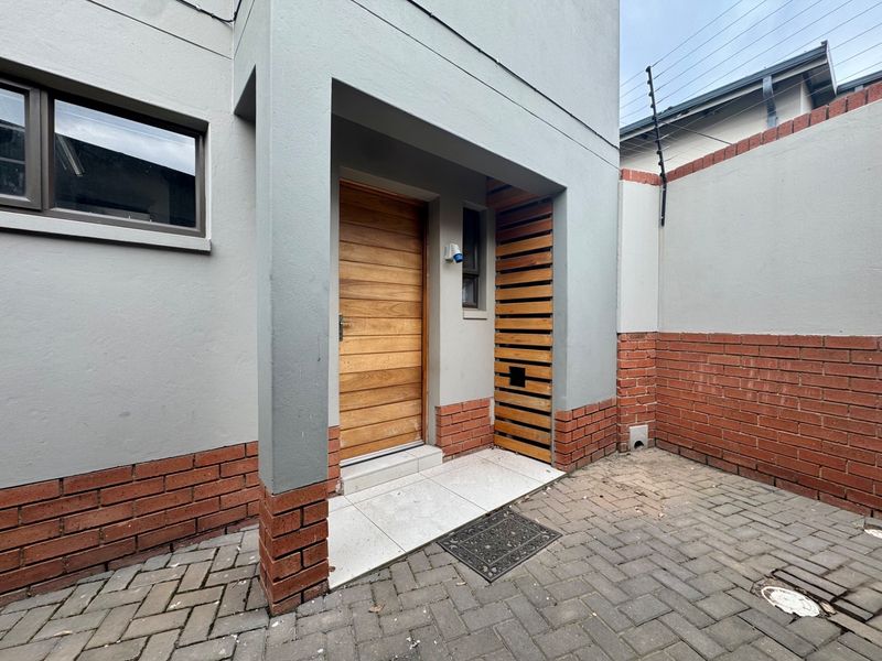 A modern townhouse entrance featuring a timber front door, decorative timber slat screen, and a paved courtyard area with secure boundary walls.