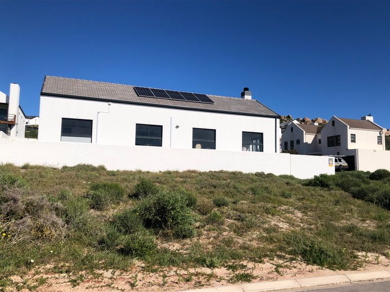 A modern residential property featuring a white facade, tiled roof with integrated solar panels, and a surrounding boundary wall on a sloped plot.