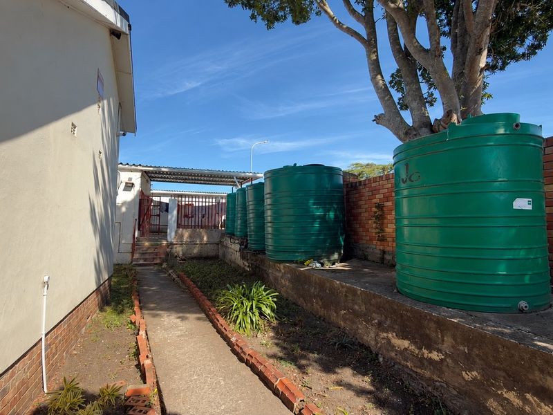Rear exterior area featuring multiple water storage tanks, a brick boundary wall, and a concrete pathway along the side of the house.