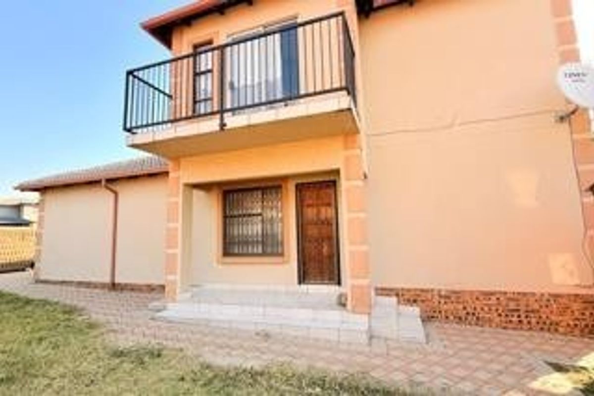 Rear view of the house featuring a paved patio area, a first-floor balcony with metal railings, and security features on windows and doors.