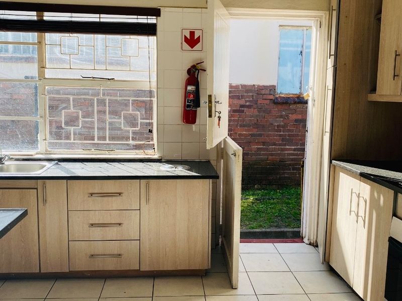 A functional kitchen featuring light wood-finish cabinetry, dark countertops, tiled flooring, and direct access to the rear exterior through a security door.