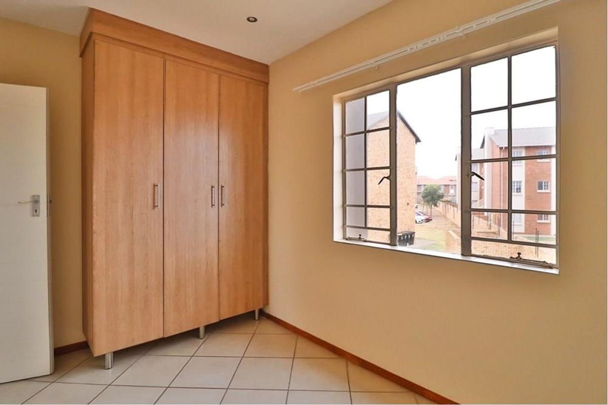 A bright, empty bedroom featuring neutral tiled flooring, a large built-in wooden wardrobe, and a window with security bars overlooking the complex.
