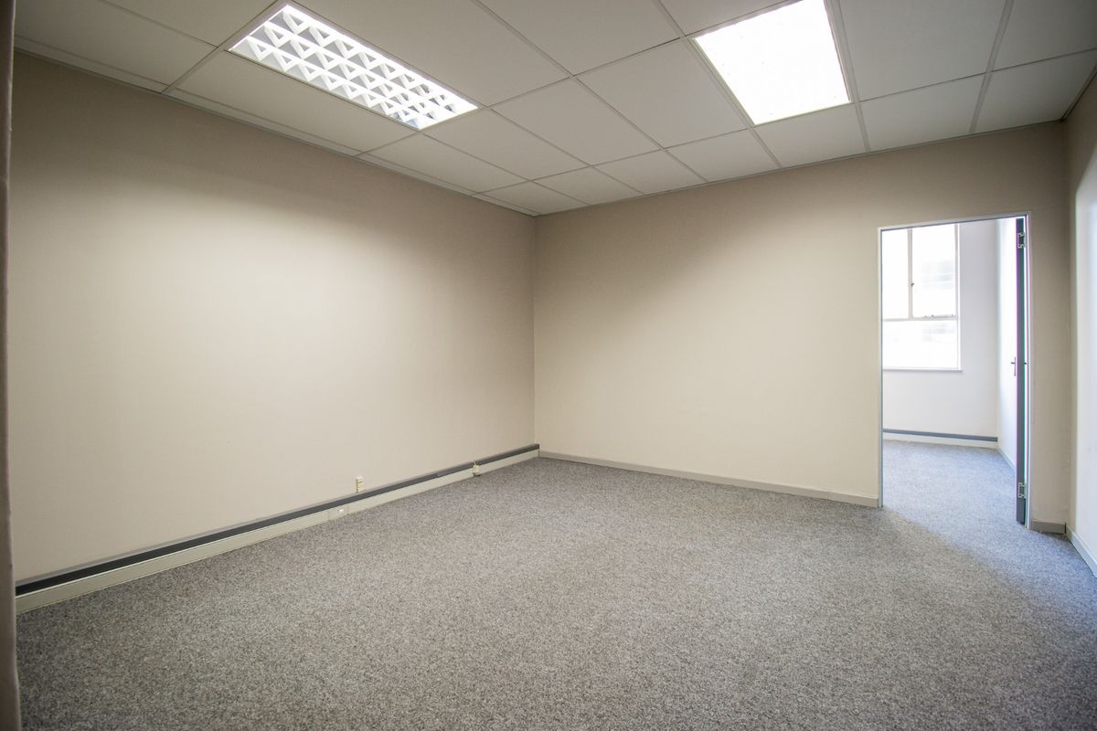 A clean, empty office space featuring neutral walls, grey carpet flooring, and a suspended ceiling with recessed lighting.
