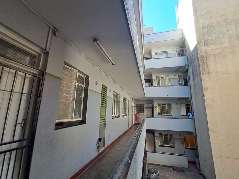 An exterior apartment building walkway featuring concrete flooring, metal security gates, and multiple window access points along the building facade.