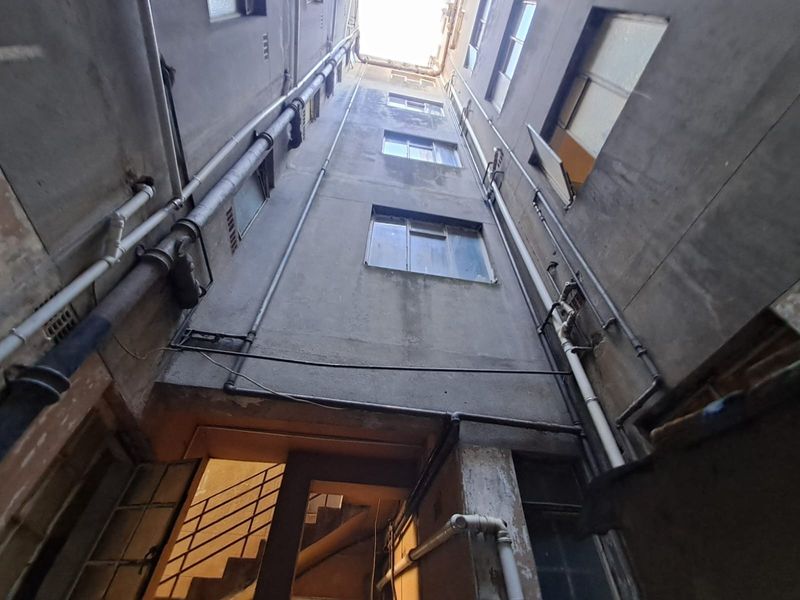A view looking up a narrow light well or rear service area of an apartment building, featuring exposed plumbing and weathered concrete walls.