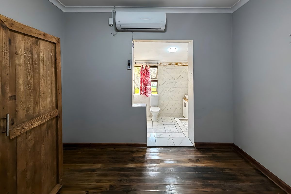 A bedroom featuring timber flooring and a wall-mounted air conditioning unit, with an open doorway leading into an adjoining bathroom.
