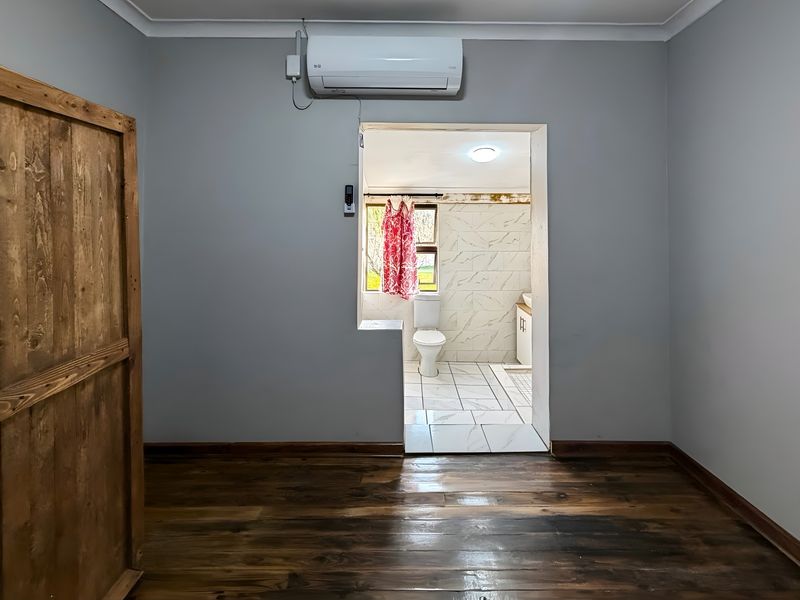A bedroom featuring timber flooring and a wall-mounted air conditioning unit, with an open doorway leading into an adjoining bathroom.