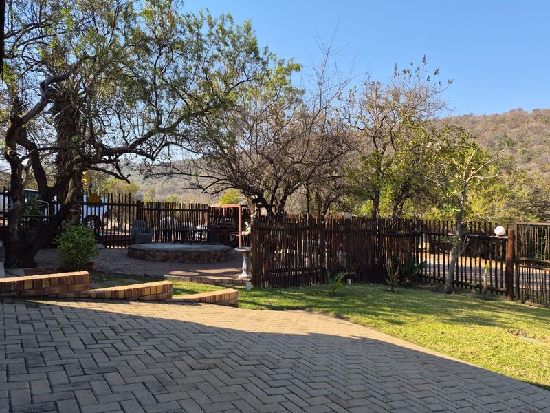 A spacious outdoor garden area featuring brick paving, a built-in fire pit, and natural timber fencing with mature trees and mountain views.