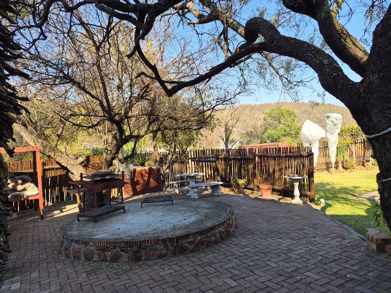 Outdoor patio area featuring brick paving, a raised circular concrete platform, and mature trees, enclosed by rustic wooden pole fencing.