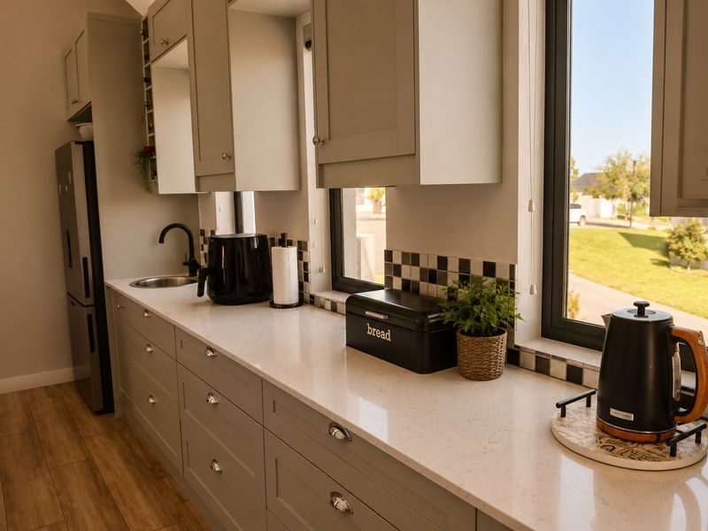 Modern kitchen featuring light grey shaker-style cabinetry, quartz countertops, a tiled backsplash, and wood-look flooring with ample natural light.