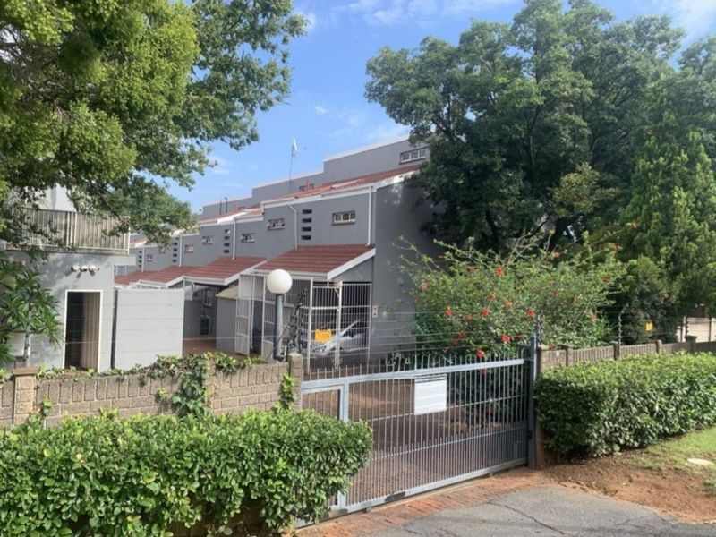 Front view of a residential complex featuring a metal security gate, brick boundary wall with electric fencing, and multi-level grey facade with tiled roofing.