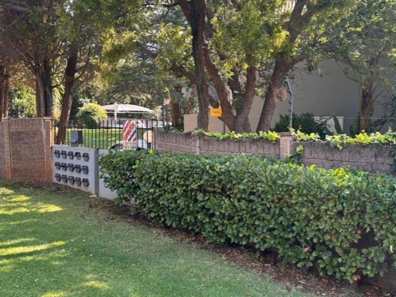 Front boundary view featuring a brick wall, metal security gate, and a bank of mailboxes set against mature trees and a landscaped hedge.