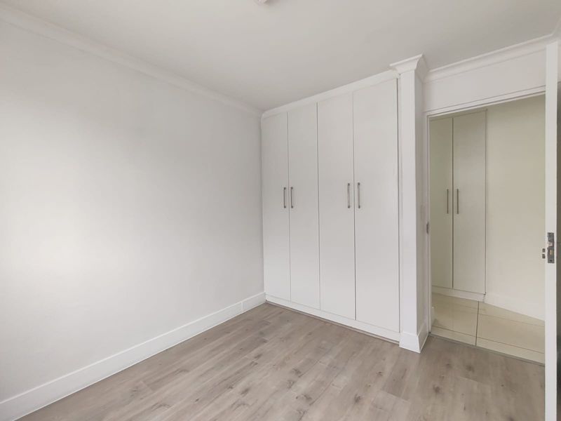 A bright, empty bedroom featuring modern laminate flooring, crisp white walls, and ample built-in cupboard storage with a clean, neutral finish.