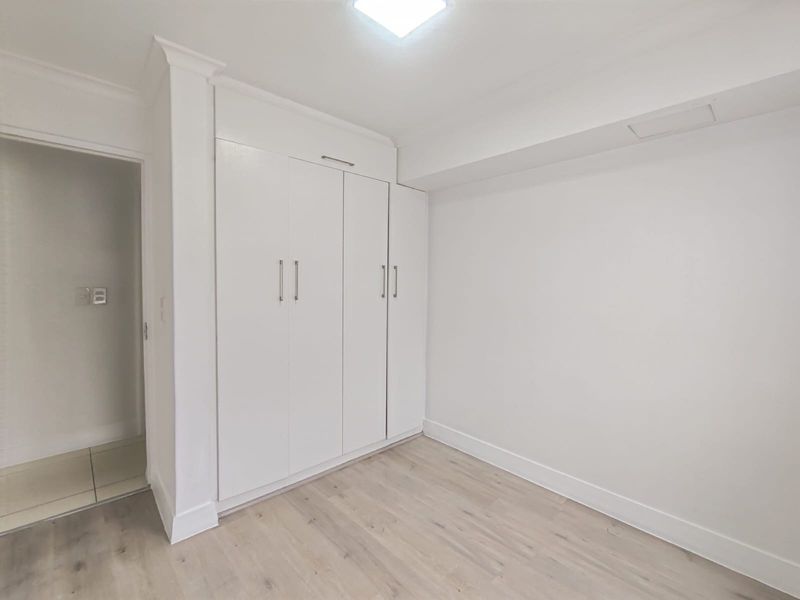 A bright, empty bedroom featuring light-toned laminate flooring and a clean, white built-in wardrobe with modern hardware.