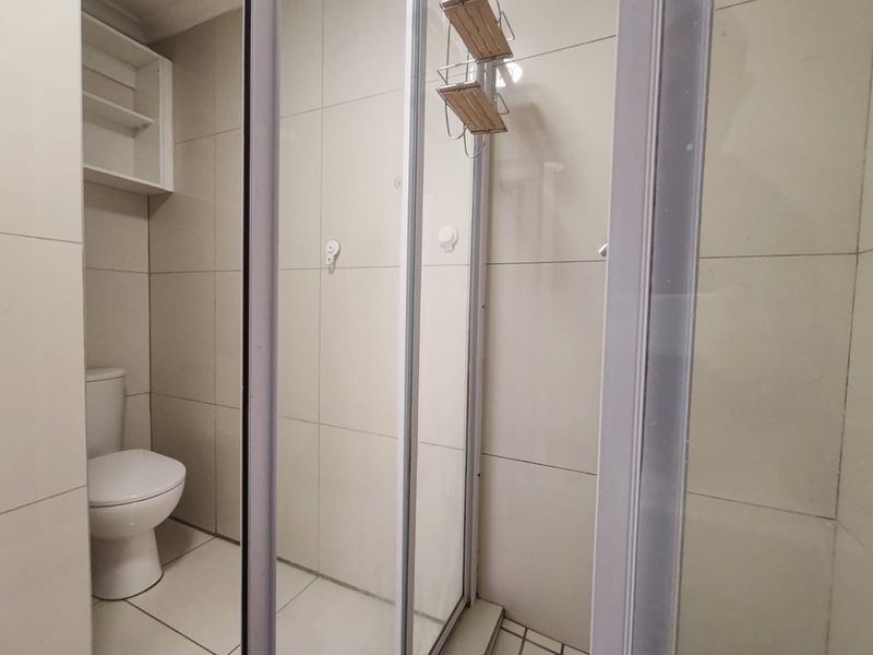 A clean, functional bathroom featuring a glass-enclosed shower, a standard toilet, and neutral wall tiling with built-in storage shelving.
