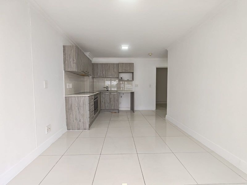 A clean, modern kitchen featuring light wood-grain cabinetry, tiled flooring, and integrated appliances, presented in an empty, well-maintained space.