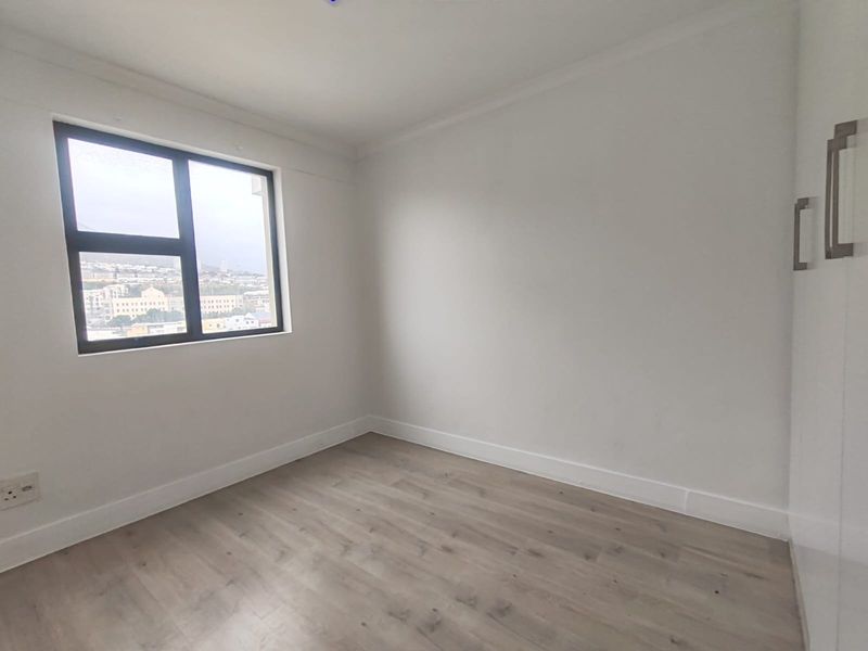 A bright, empty bedroom featuring modern laminate flooring, a built-in wardrobe, and a window with an urban view.