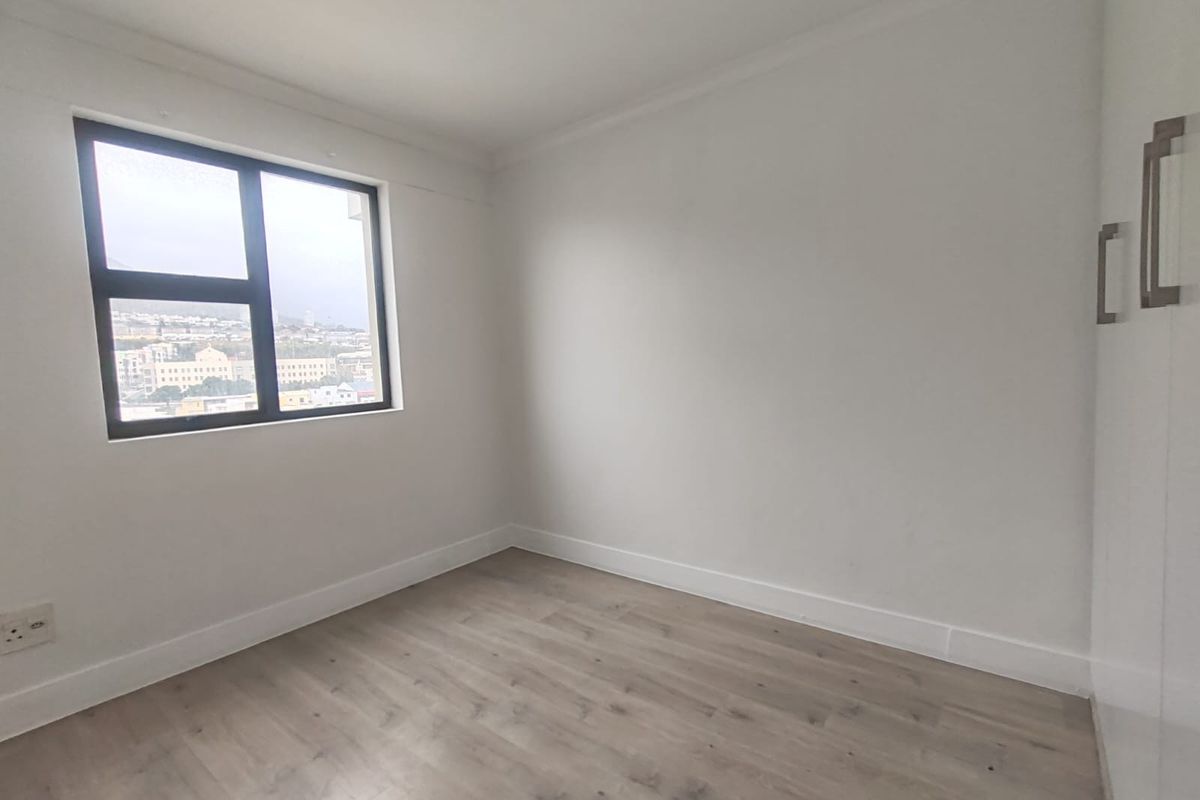 A bright, empty bedroom featuring modern laminate flooring, a built-in wardrobe, and a window with an urban view.