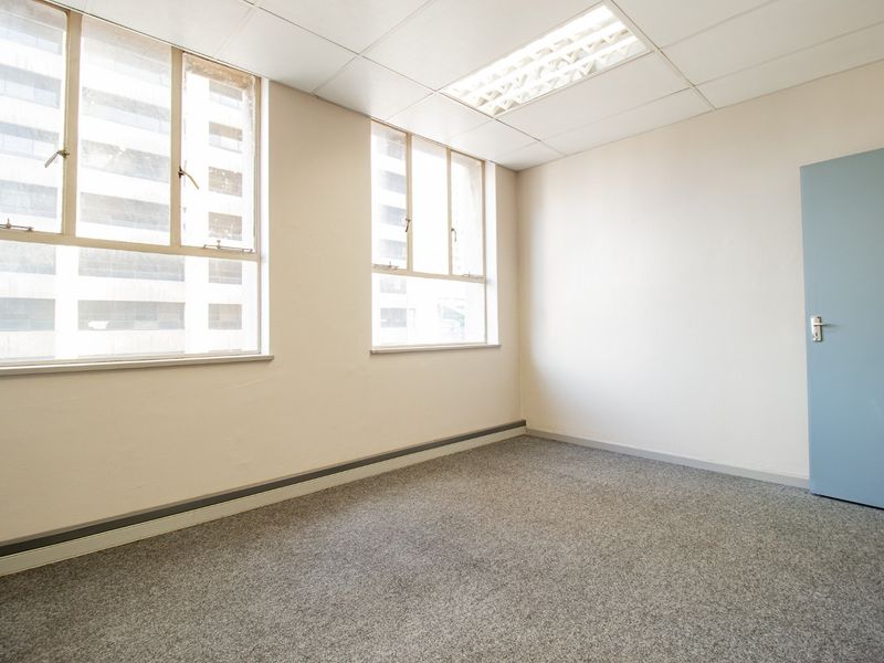 A vacant commercial office space featuring neutral walls, carpeted flooring, suspended ceiling with recessed lighting, and large windows providing natural light.