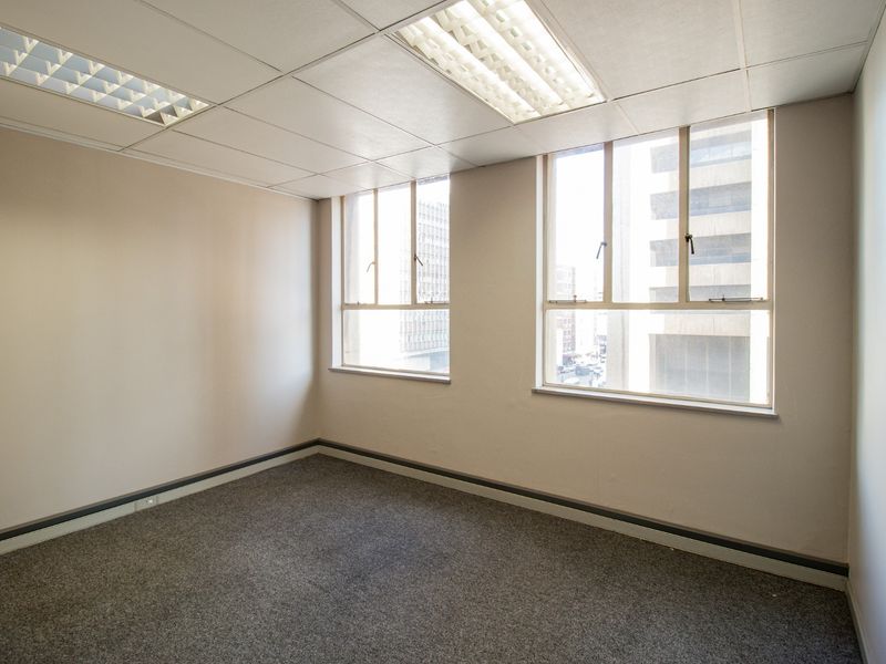 A vacant office space featuring neutral walls, grey carpeting, suspended ceiling tiles with recessed lighting, and large windows providing natural light.