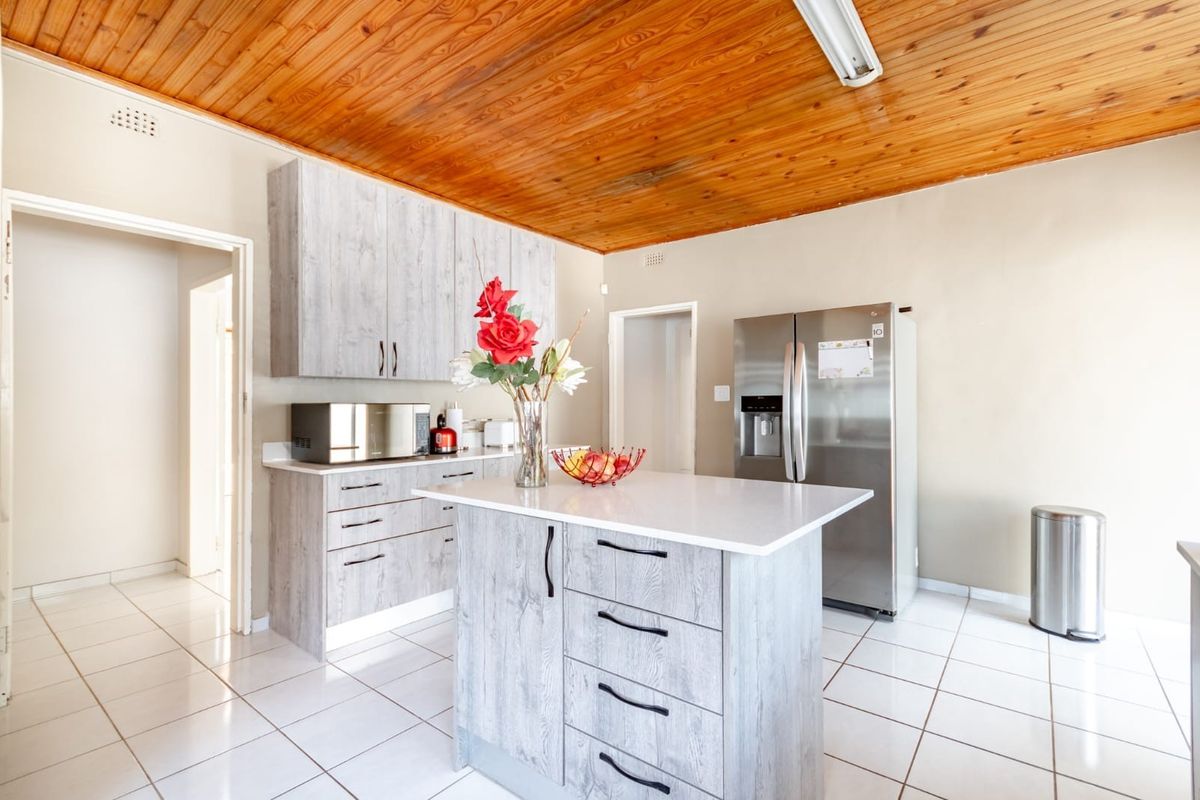 A functional kitchen featuring modern grey cabinetry, a central island with stone countertop, tiled flooring, and a distinctive timber-clad ceiling.