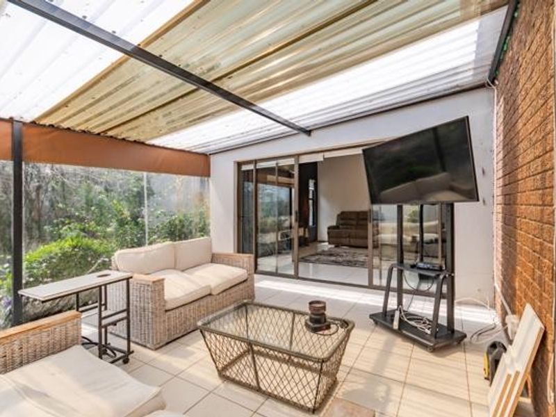 A covered patio area featuring tiled flooring, an exposed brick wall, and a translucent roof, providing a functional outdoor living space.
