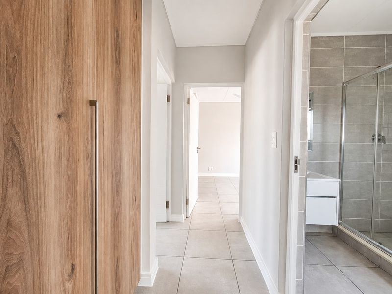 A clean, modern hallway featuring light grey floor tiles, wood-finish built-in cupboards, and access to adjacent rooms including a bathroom.