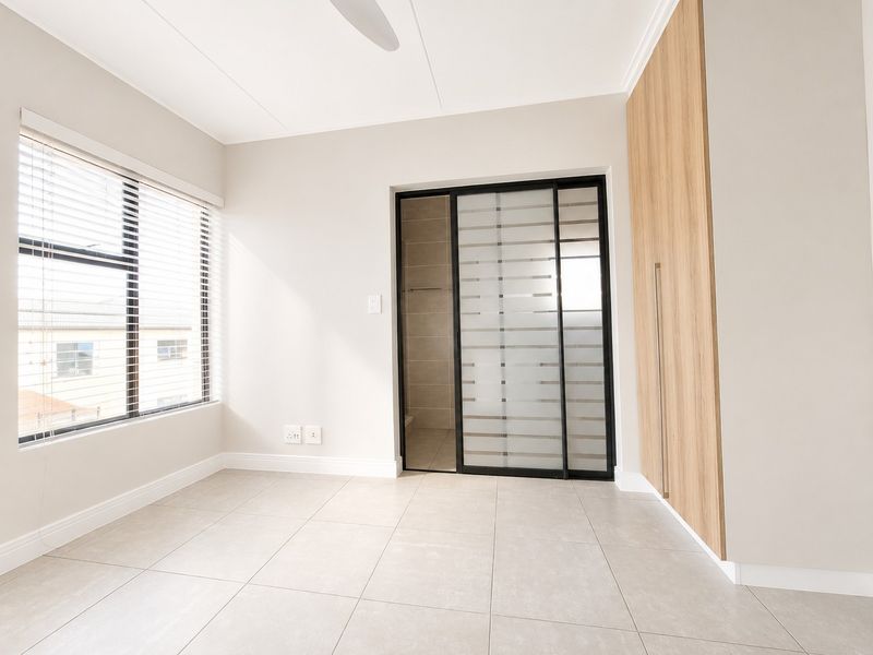 A bright, modern bedroom featuring neutral tiled flooring, built-in wooden cupboards, and a sleek sliding glass door leading to an en-suite bathroom.