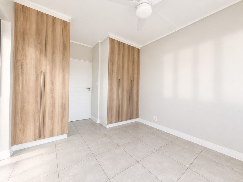 A bright, empty bedroom featuring modern wood-finish built-in cupboards, neutral tiled flooring, and a ceiling fan.