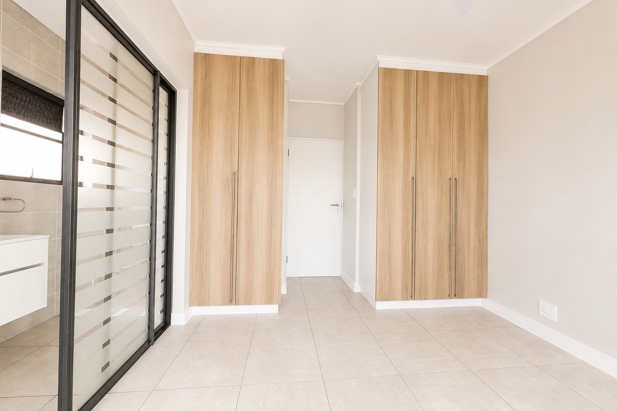 A bright, modern bedroom featuring light wood-finish built-in cupboards, neutral floor tiles, and a sleek glass sliding door leading to an en-suite bathroom.