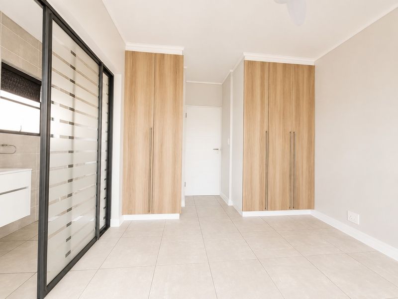 A bright, modern bedroom featuring light wood-finish built-in cupboards, neutral floor tiles, and a sleek glass sliding door leading to an en-suite bathroom.