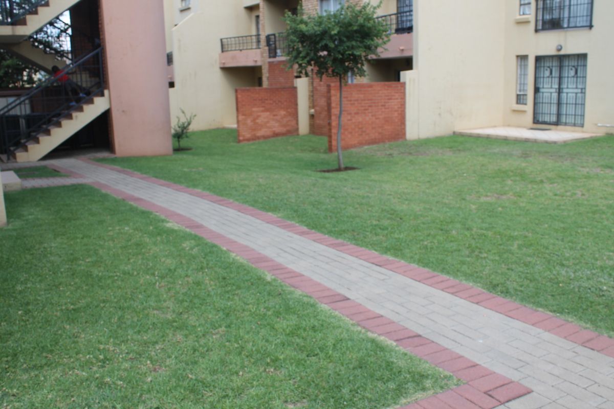Communal garden area featuring a paved walkway, brick boundary walls, and access to apartment building staircases and entrances.