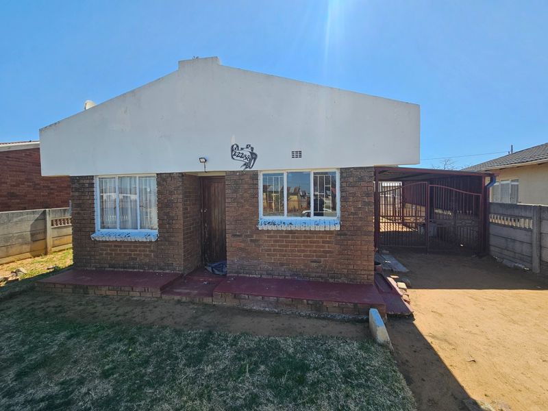 Front elevation of a single-story brick house featuring a carport, metal security gate, and a small front yard with a concrete boundary wall.