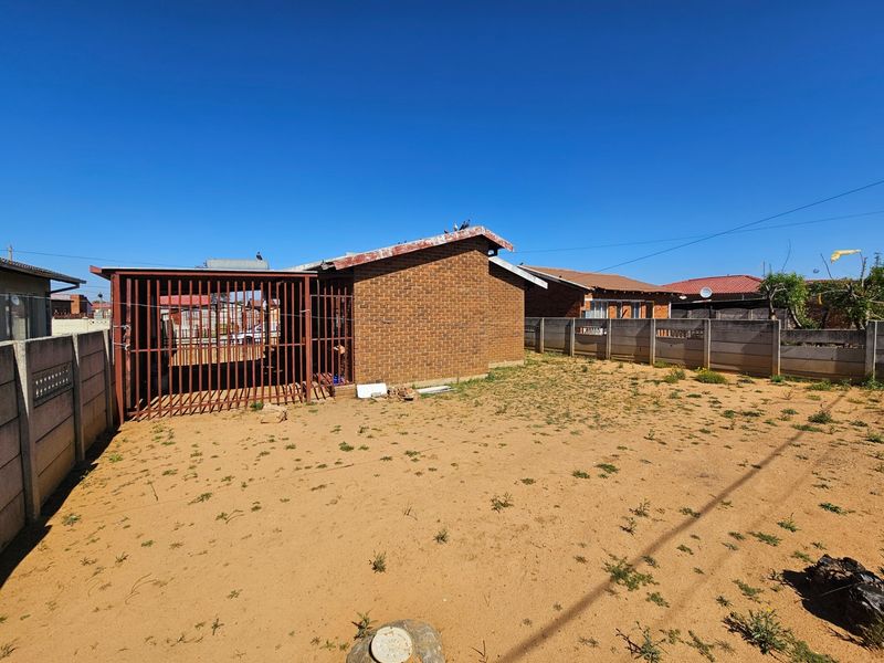Rear view of a face brick house featuring a large sandy yard, enclosed by precast concrete boundary walls and a metal security gate.