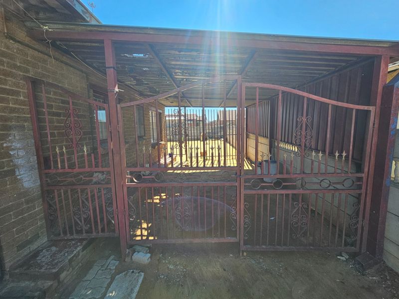 A rear exterior area featuring a covered patio space enclosed by a metal security gate, showing signs of wear and requiring maintenance.