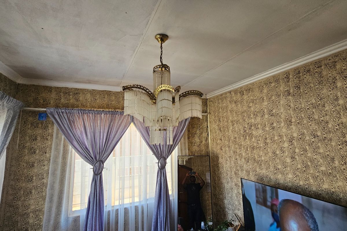 A living room featuring a prominent crystal chandelier, decorative ceiling cornice, and patterned wallpaper, requiring cosmetic updates to modernize the space.