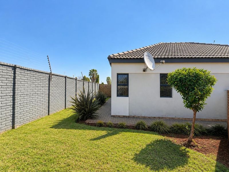 A well-maintained rear exterior view featuring a paved walkway, manicured lawn, and secure brick boundary wall with electric fencing.