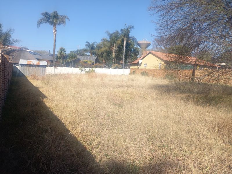 A vacant residential plot featuring dry, overgrown grass, a perimeter boundary wall, and a small metal storage shed.