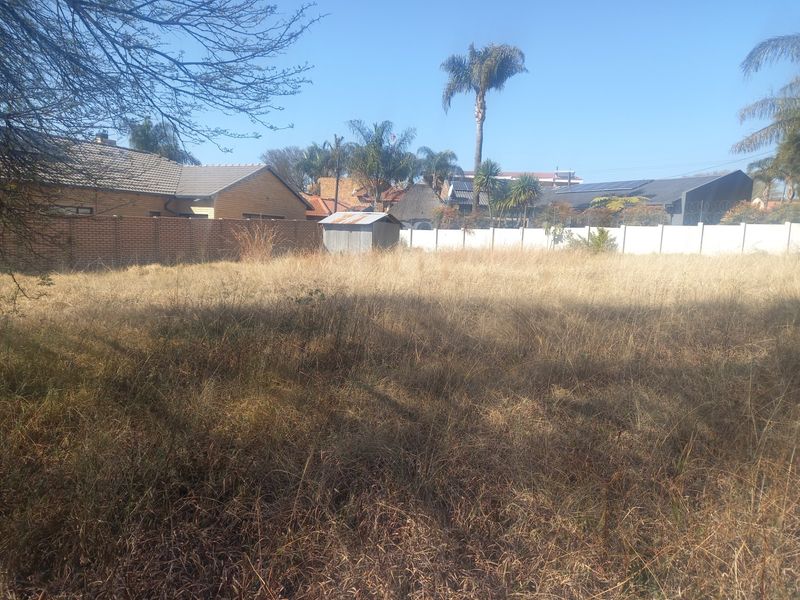 A vacant residential plot featuring dry, overgrown grass, enclosed by brick and precast concrete boundary walls with a small metal storage shed.
