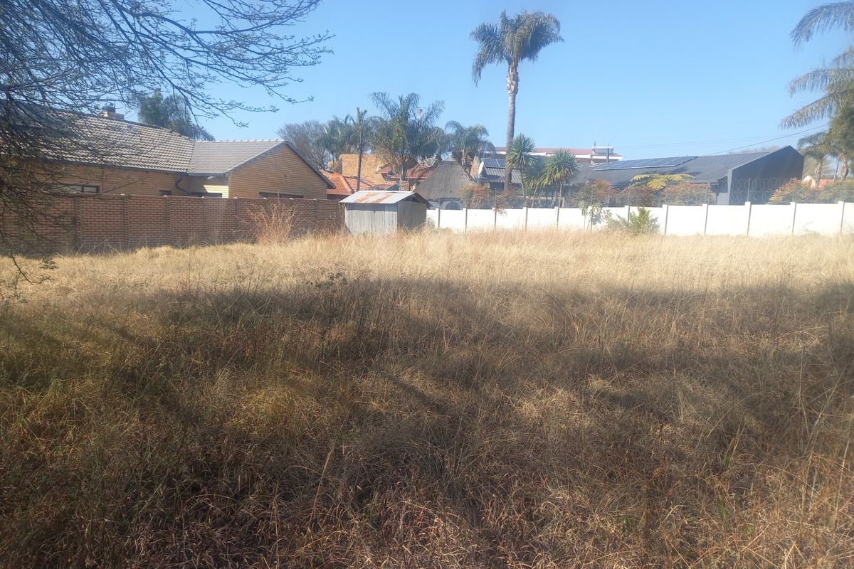 A vacant residential plot featuring dry, overgrown grass, enclosed by brick and precast concrete boundary walls with a small metal storage shed.