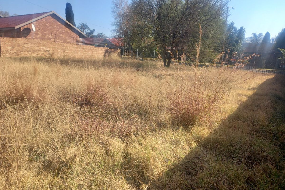 A vacant residential plot featuring overgrown grass and established trees, enclosed by a brick boundary wall and perimeter fencing.