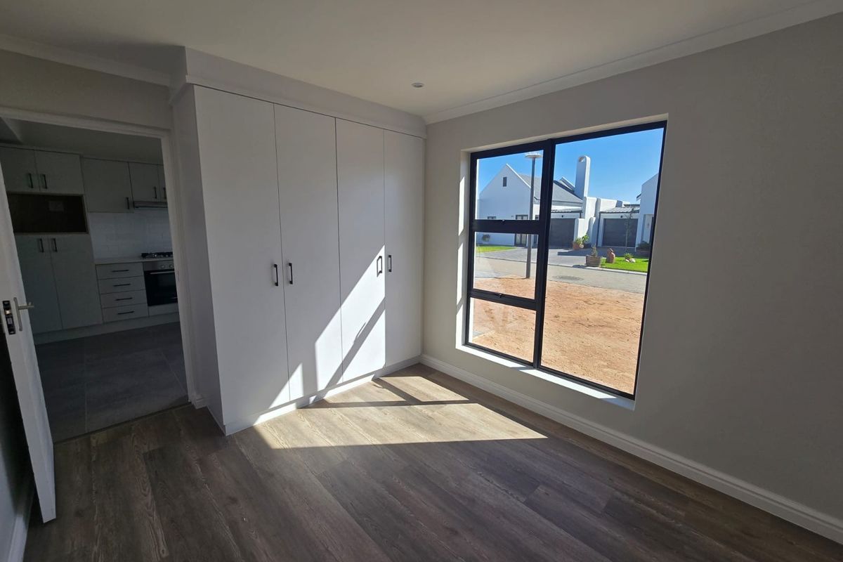 A bright, modern bedroom featuring neutral walls, durable laminate flooring, and ample built-in cupboard space, with a view towards the street.