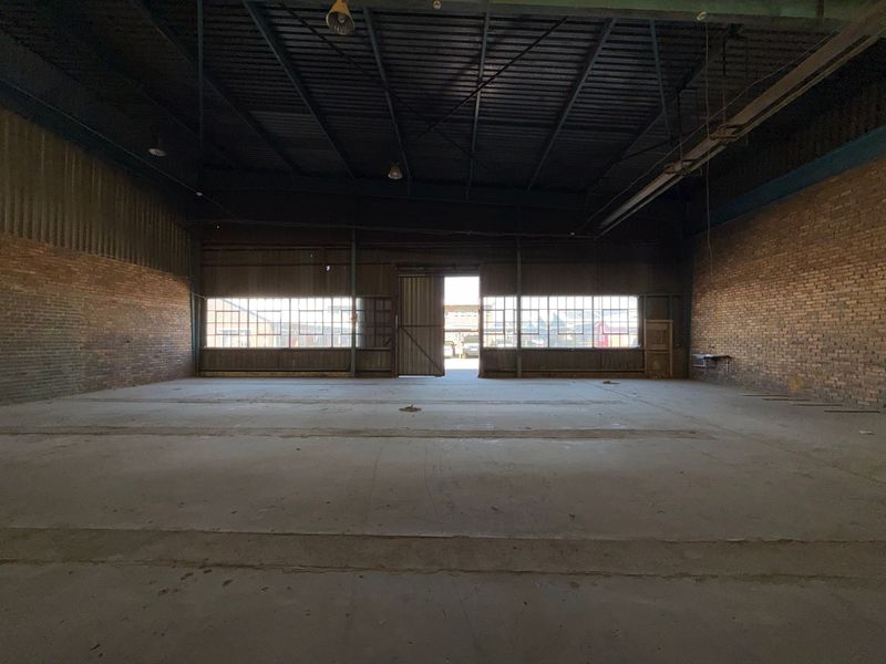 Spacious industrial warehouse unit featuring exposed brick walls, concrete flooring, and high-clearance steel roof structure with natural light from large metal-framed windows.