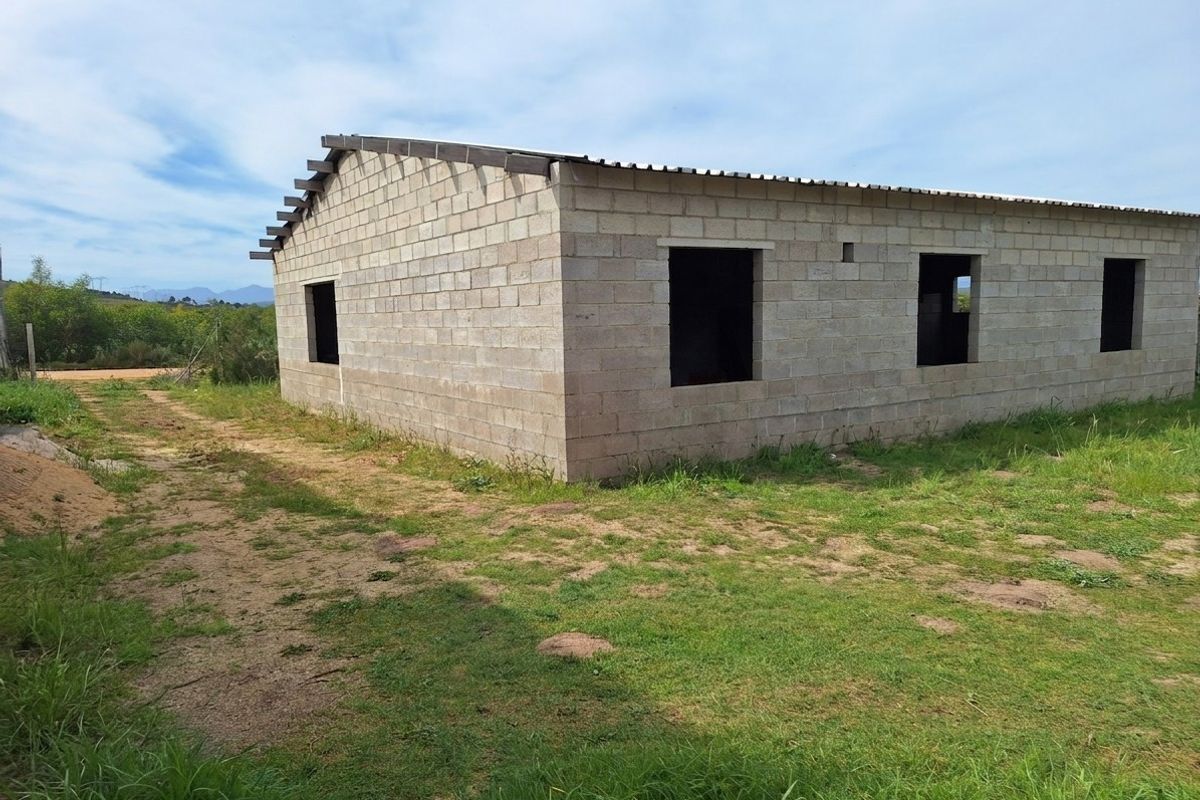 An unfinished residential structure featuring concrete block walls and a corrugated metal roof, set on a vacant plot of land.