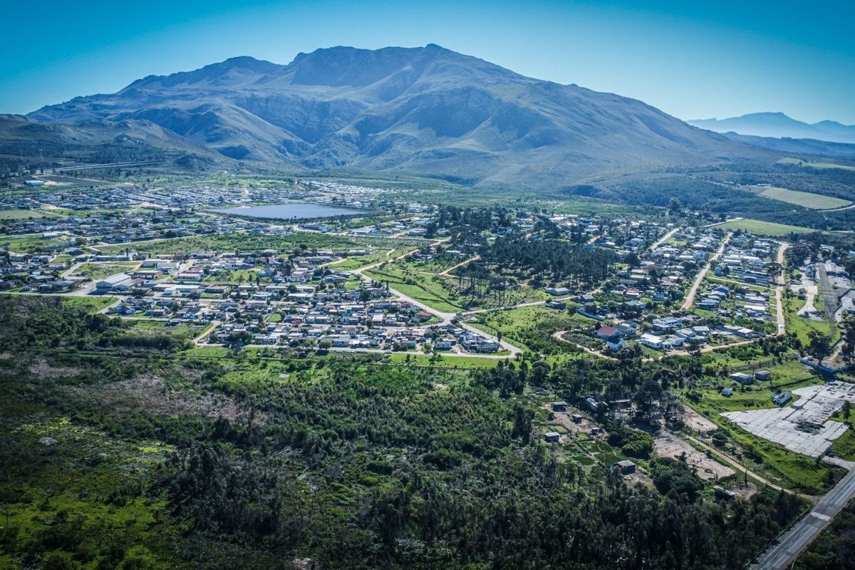 An aerial view of a residential township situated at the base of a large mountain range, showcasing the surrounding landscape and road infrastructure.