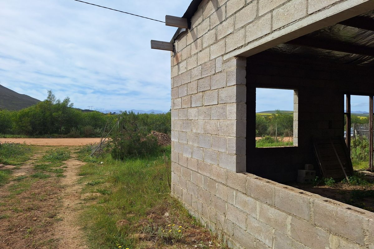 An unfinished residential structure featuring exposed concrete block walls and timber rafters, situated on a vacant plot with surrounding landscape views.