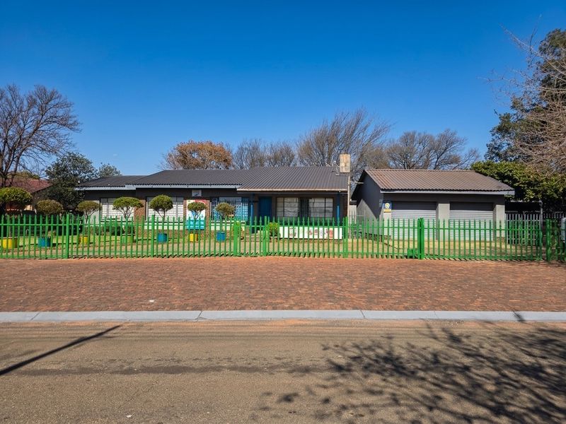 Front view of a single-story house featuring a brick facade, pitched metal roof, double garage, and secure metal perimeter fencing.
