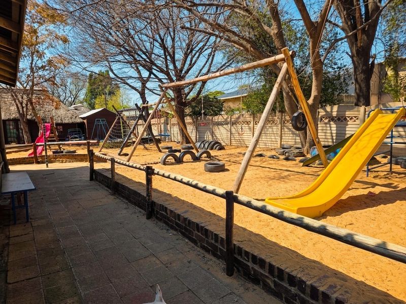 A spacious outdoor play area featuring a sand pit, paved walkway, and established trees, enclosed by a combination of timber and precast concrete boundary walls.