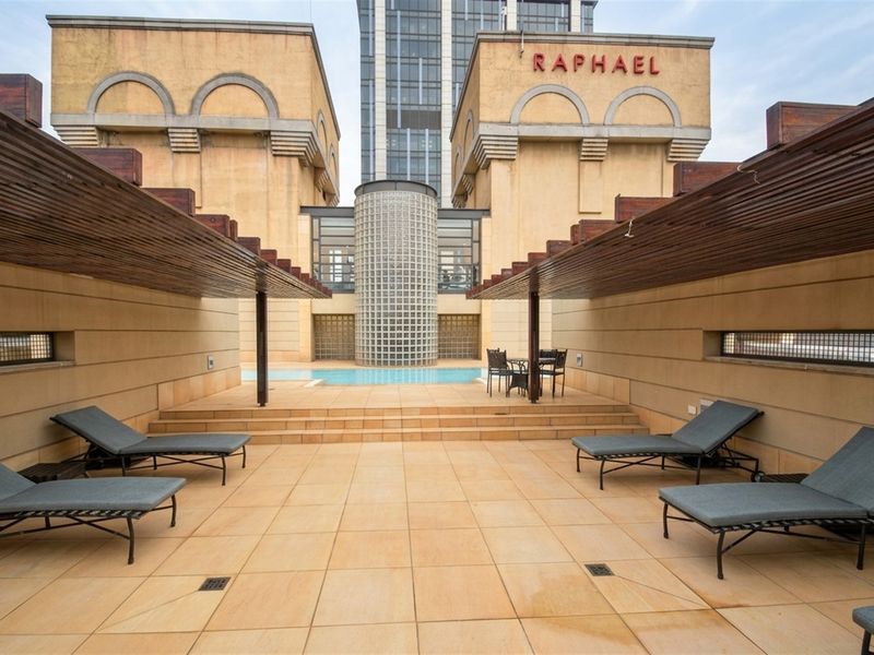A communal pool area featuring a central swimming pool, glass block architectural detail, and a spacious tiled terrace with timber pergola structures.