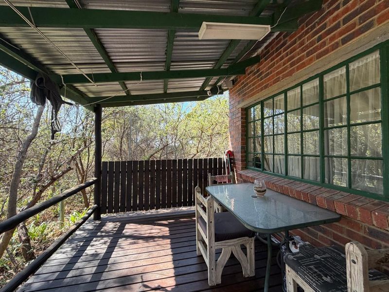 A covered outdoor patio featuring timber decking, exposed brickwork, and a corrugated metal roof, overlooking a natural wooded area.